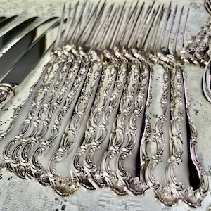 May include: An assortment of ornate silver-colored flatware, including knives, forks, and spoons. The handles feature a detailed, scrolling design. The silverware is arranged on a white, textured surface, suggesting a formal dining setting.