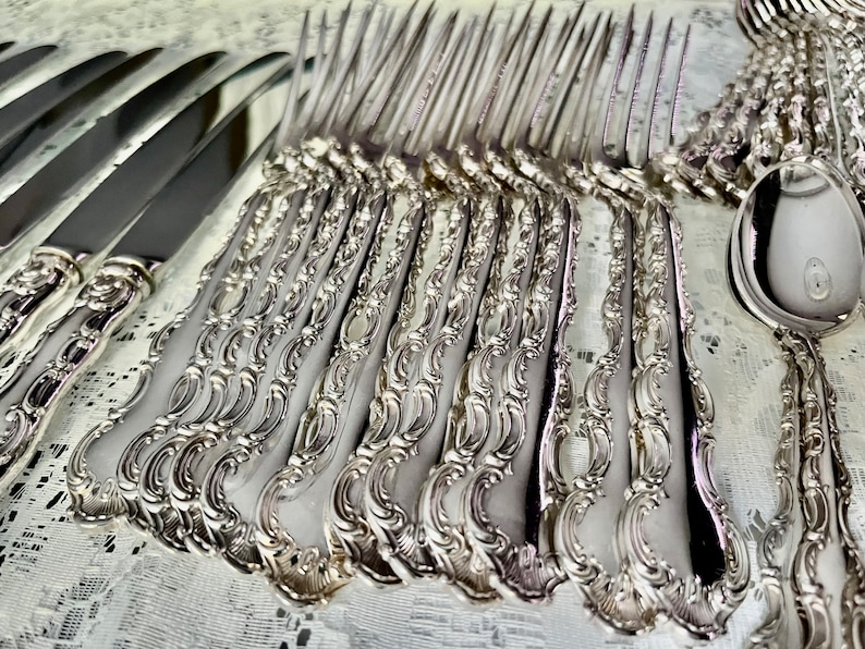 May include: An assortment of ornate silver-colored flatware, including knives, forks, and spoons. The handles feature a detailed, scrolling design. The silverware is arranged on a white, textured surface, suggesting a formal dining setting.