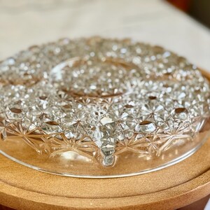 May include: A clear, cut-glass serving dish with a starburst pattern, resting on a round wooden tray. The dish has a central indentation and a decorative, textured surface, perfect for serving appetizers or desserts.