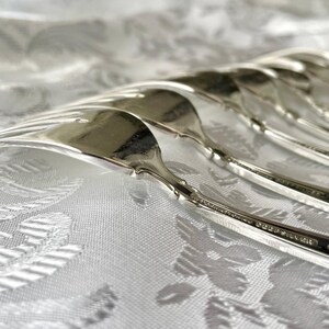 May include: Close-up of a set of silver forks arranged on a white patterned tablecloth. The forks are highly polished, reflecting light, and have a classic, elegant design. The image highlights the silverware's details and the texture of the fabric.