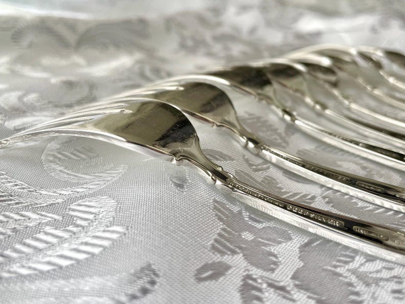 May include: Close-up of a set of silver forks arranged on a white patterned tablecloth. The forks are highly polished, reflecting light, and have a classic, elegant design. The image highlights the silverware's details and the texture of the fabric.