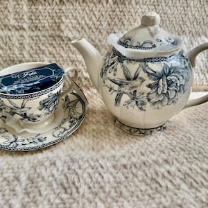 K&ouml;nnte beinhalten: Ein wei&szlig;es Porzellan-Teeservice mit einer passenden Teekanne. Das Set hat ein blaues Blumen- und Vogeldesign. Die Teekanne hat einen grauen Griff und Deckel. Die Tasse hat eine passende Untertasse und ein Etikett.