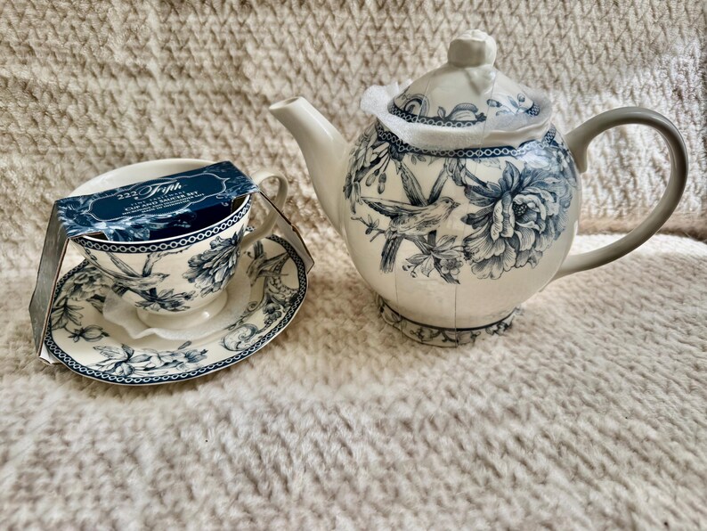 K&ouml;nnte beinhalten: Ein wei&szlig;es Porzellan-Teeservice mit einer passenden Teekanne. Das Set hat ein blaues Blumen- und Vogeldesign. Die Teekanne hat einen grauen Griff und Deckel. Die Tasse hat eine passende Untertasse und ein Etikett.