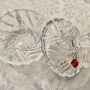 May include: A clear, cut-glass candy dish with a matching lid. The dish features a radial, starburst-like pattern of deep, faceted grooves. The lid includes a faceted handle and a small, red, circular sticker with gold text attached to the surface.