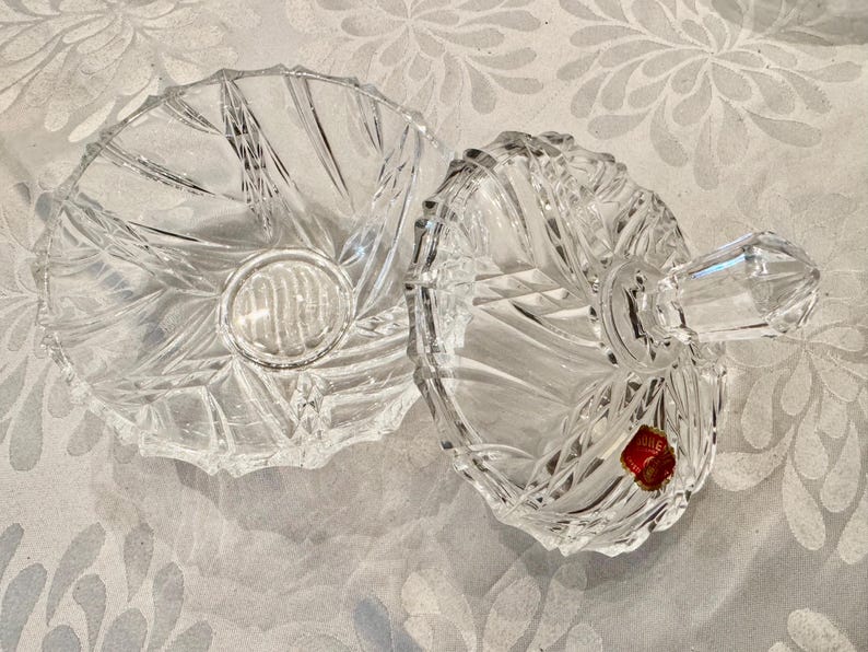 May include: A clear, cut-glass candy dish with a matching lid. The dish features a radial, starburst-like pattern of deep, faceted grooves. The lid includes a faceted handle and a small, red, circular sticker with gold text attached to the surface.