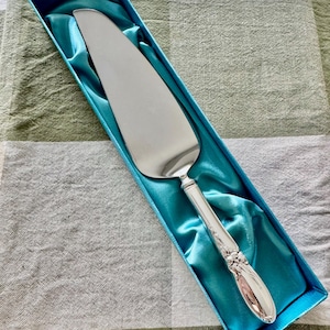 Op de afbeelding: Een zilveren taartschep met een decoratief handvat, gepresenteerd in een blauwe geschenkdoos gevoerd met satijnen stof. De schep heeft een breed, plat lemmet en sierlijke details op het handvat. De doos is rechthoekig.
