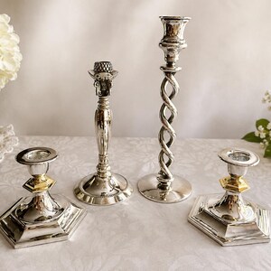 May include: A set of four silver-toned metal candle holders. Two feature hexagonal bases with gold-toned accents, one has a smooth tapered stem with a textured top, and the tallest one displays a twisted spiral stem design with a circular base.