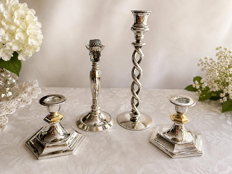May include: A set of four silver-toned metal candle holders. Two feature hexagonal bases with gold-toned accents, one has a smooth tapered stem with a textured top, and the tallest one displays a twisted spiral stem design with a circular base.