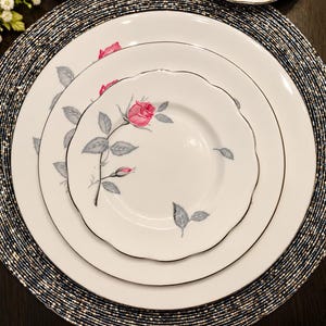 May include: A set of three stacked white porcelain plates with a silver rim. Each plate features a delicate illustration of a pink rosebud with gray leaves and stems. The smallest plate has a scalloped edge, while the larger plates are round.