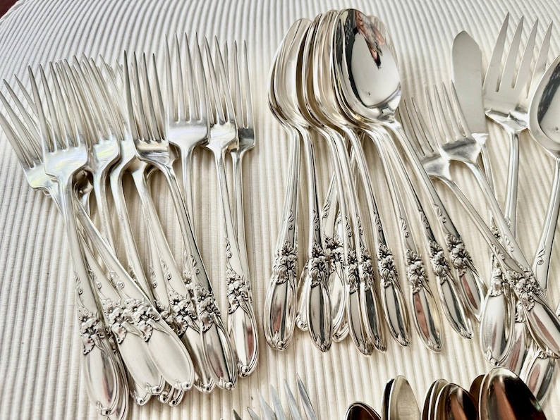 Op de afbeelding: Een verzameling antiek zilverwerk, waaronder vorken, lepels en messen, gerangschikt op een gestructureerd oppervlak. Het zilveren bestek heeft bloemmotieven op de handvatten. De lepels en vorken zijn in nette rijen gerangschikt.