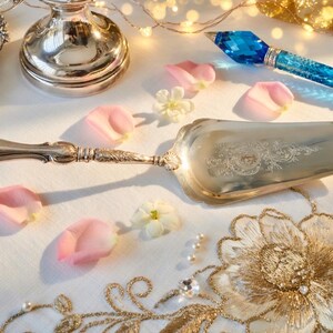 K&ouml;nnte beinhalten: Ein silberner Tortenheber mit einem verzierten Griff und floralem Design auf der Klinge. Der Heber ist von rosa Rosenbl&auml;ttern, kleinen wei&szlig;en Bl&uuml;ten und einem blauen Kristallstift umgeben. Eine silberne Servierschale und ein goldfarbenes Blumenmuster sind ebenfalls zu sehen.