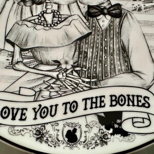 May include: A black and white decorative plate featuring a skeleton couple. The plate has the phrase "LOVE YOU TO THE BONES" on a banner. The design includes floral and skull motifs, and musical notes.