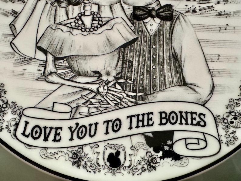 May include: A black and white decorative plate featuring a skeleton couple. The plate has the phrase "LOVE YOU TO THE BONES" on a banner. The design includes floral and skull motifs, and musical notes.