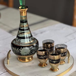 May include: A heart-shaped marble serving tray with a gold rim and handle. The tray holds a green and gold glass decanter and four matching shot glasses with black and gold designs.