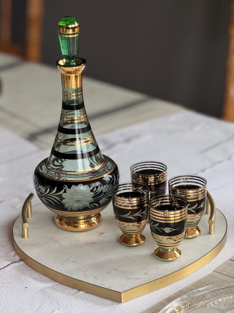 May include: A heart-shaped marble serving tray with a gold rim and handle. The tray holds a green and gold glass decanter and four matching shot glasses with black and gold designs.