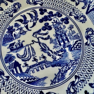 K&ouml;nnte beinhalten: Wei&szlig;e Keramikplatte mit blauem Willow-Muster. Die Platte zeigt eine detaillierte Landschaftsszene mit einer Pagode, einer Br&uuml;cke und Figuren, umgeben von dekorativen Bord&uuml;ren. Ein klassisches St&uuml;ck zum Servieren oder Ausstellen.