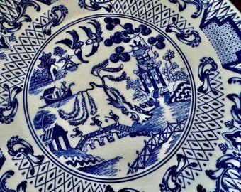 May include: White ceramic plate with a blue Willow pattern design. The plate features a detailed landscape scene with a pagoda, bridge, and figures, surrounded by decorative borders. A classic piece for serving or display.
