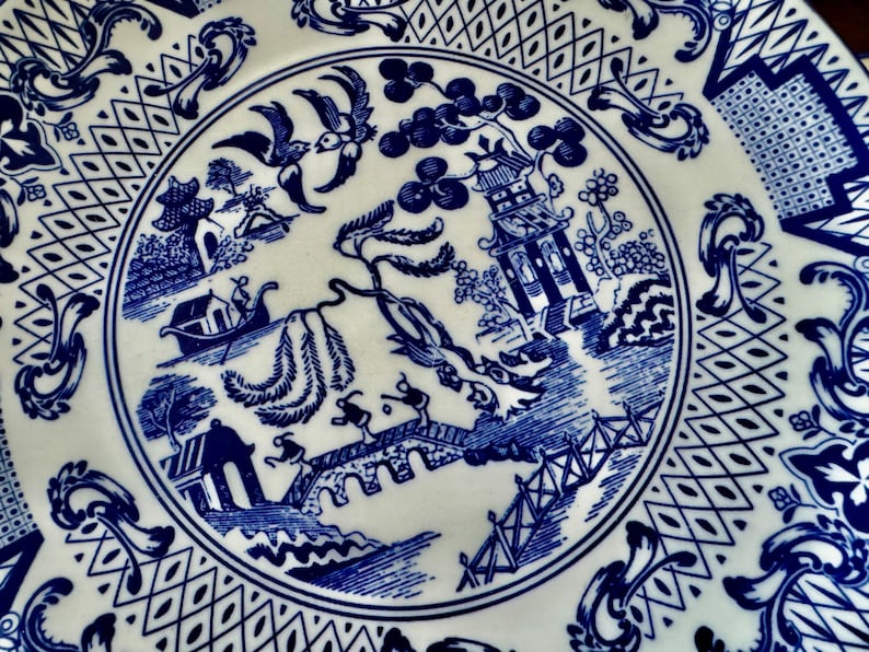 K&ouml;nnte beinhalten: Wei&szlig;e Keramikplatte mit blauem Willow-Muster. Die Platte zeigt eine detaillierte Landschaftsszene mit einer Pagode, einer Br&uuml;cke und Figuren, umgeben von dekorativen Bord&uuml;ren. Ein klassisches St&uuml;ck zum Servieren oder Ausstellen.