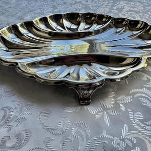 Pode incluir: Uma bandeja de servir em metal prateado com formato de uma grande concha de vieira. A bandeja apresenta uma borda recortada, uma superf&iacute;cie lisa e reflexiva com sulcos radiais em relevo, apoiada por um &uacute;nico p&eacute; de metal decorativo.
