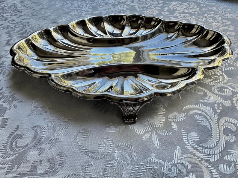 Pode incluir: Uma bandeja de servir em metal prateado com formato de uma grande concha de vieira. A bandeja apresenta uma borda recortada, uma superf&iacute;cie lisa e reflexiva com sulcos radiais em relevo, apoiada por um &uacute;nico p&eacute; de metal decorativo.