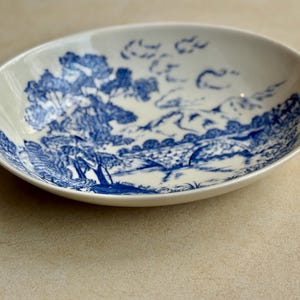 May include: Oval, off-white ceramic dish with a blue landscape design. The design features trees, a bridge, and mountains. The dish is likely for serving food or as a decorative piece.