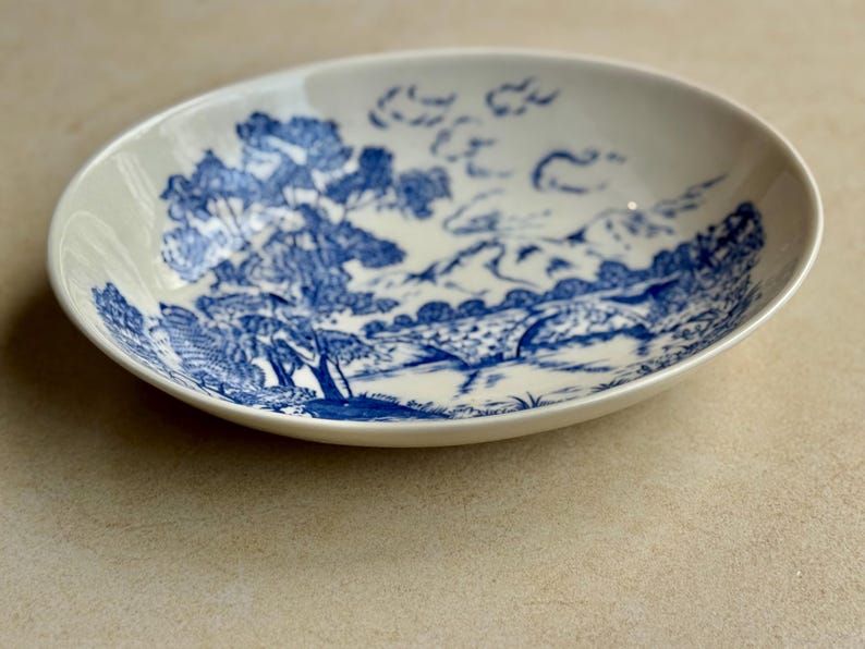 May include: Oval, off-white ceramic dish with a blue landscape design. The design features trees, a bridge, and mountains. The dish is likely for serving food or as a decorative piece.