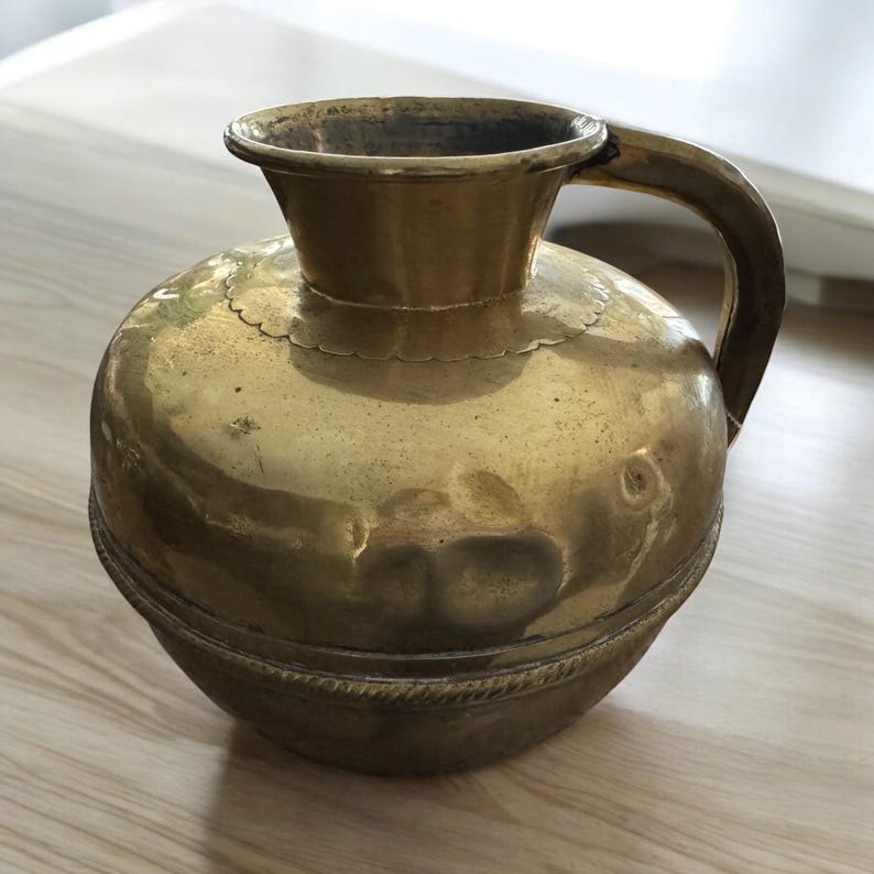 K&ouml;nnte beinhalten: Eine goldfarbene Messingkanne mit rundem K&ouml;rper und Henkel. Die Kanne hat ein dekoratives Band um die Mitte.