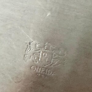 May include: A silver-toned metal surface featuring an embossed maker's mark. The stamp displays a heraldic crest design with the text ONEIDA positioned directly below the shield, showing clear metallic texture and light reflections.
