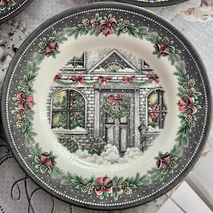 May include: A decorative plate with a Christmas scene. The plate features a detailed illustration of a snow-covered house with a wreath on the door and festive decorations. The rim is dark gray with a dotted pattern and floral accents.