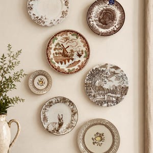 May include: A collection of six decorative ceramic plates mounted on a wall, featuring various brown and white transferware patterns including a turkey, a windmill, a rabbit, and floral motifs, alongside a cream-colored ceramic pitcher.