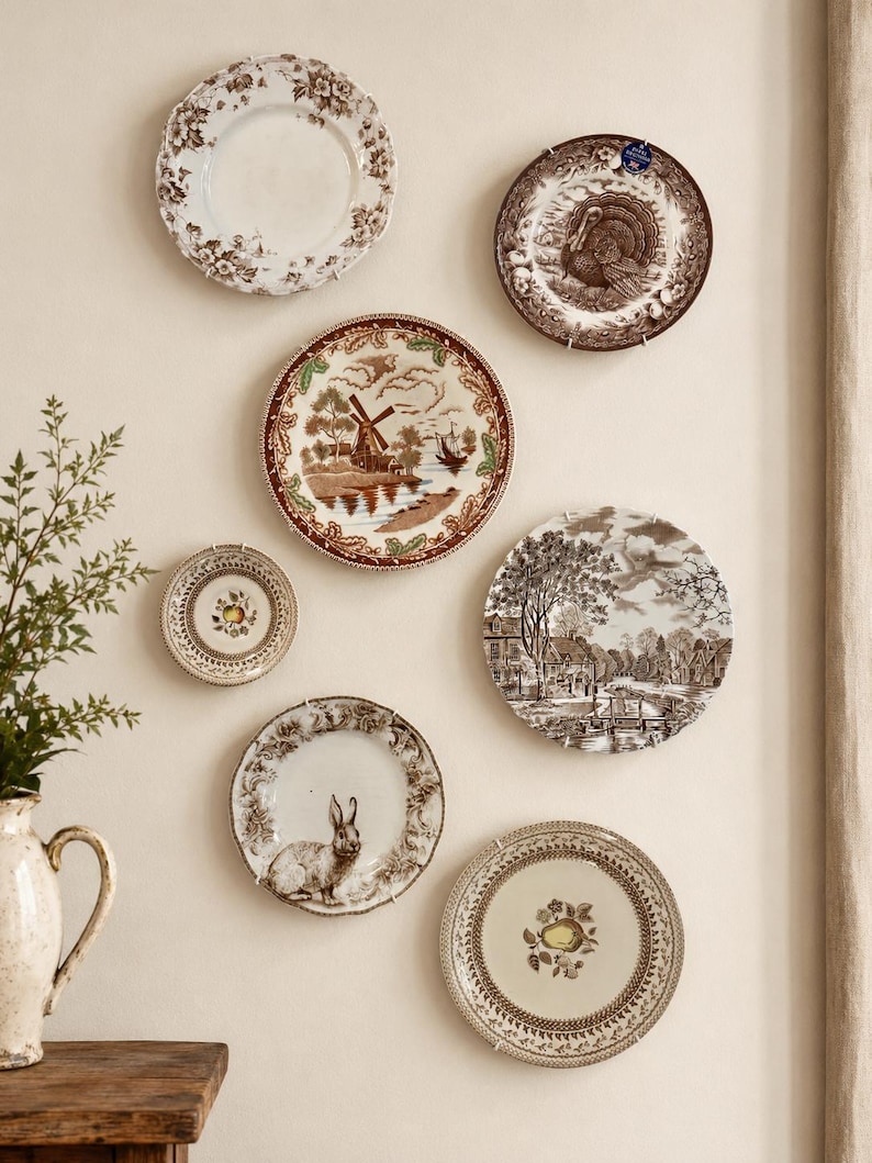 May include: A collection of six decorative ceramic plates mounted on a wall, featuring various brown and white transferware patterns including a turkey, a windmill, a rabbit, and floral motifs, alongside a cream-colored ceramic pitcher.