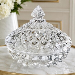 May include: A clear glass candy dish with a matching lid, featuring a faceted diamond-cut pattern across the surface. The lid is topped with a pointed finial, and the bowl has a rounded base, creating a reflective and textured appearance.