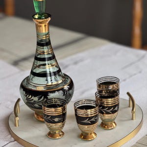 May include: A heart-shaped marble serving tray with a gold rim and handles. The tray holds a green and gold glass decanter and four matching shot glasses with black and gold designs.
