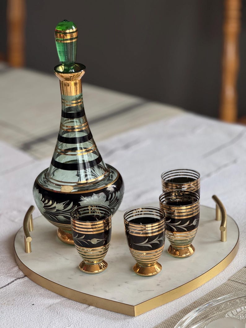 May include: A heart-shaped marble serving tray with a gold rim and handles. The tray holds a green and gold glass decanter and four matching shot glasses with black and gold designs.