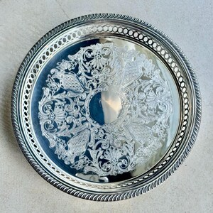 May include: A round silver-toned serving tray featuring an intricate embossed floral and scrollwork pattern in the center. The rim has a decorative pierced lattice border with a twisted rope edge design, creating a polished metallic finish.