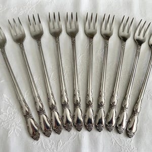 May include: A collection of ten silver forks with ornate floral detailing on the handles. The forks are arranged on a white, textured surface, showcasing their elegant design. Ideal for formal dining or special occasions.