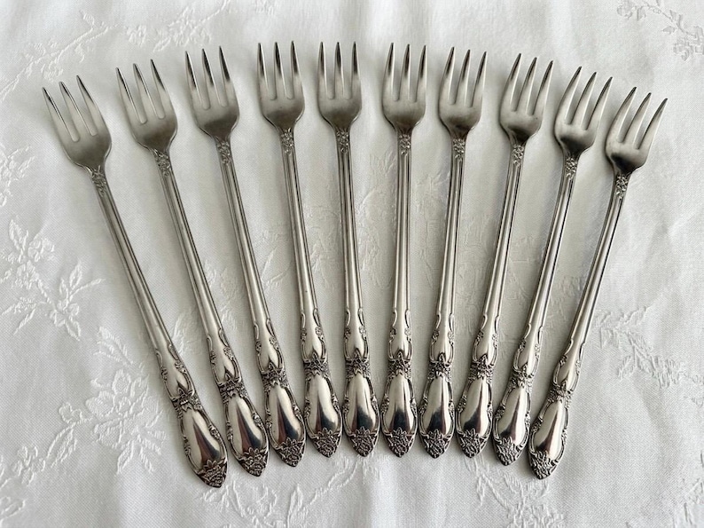May include: A collection of ten silver forks with ornate floral detailing on the handles. The forks are arranged on a white, textured surface, showcasing their elegant design. Ideal for formal dining or special occasions.