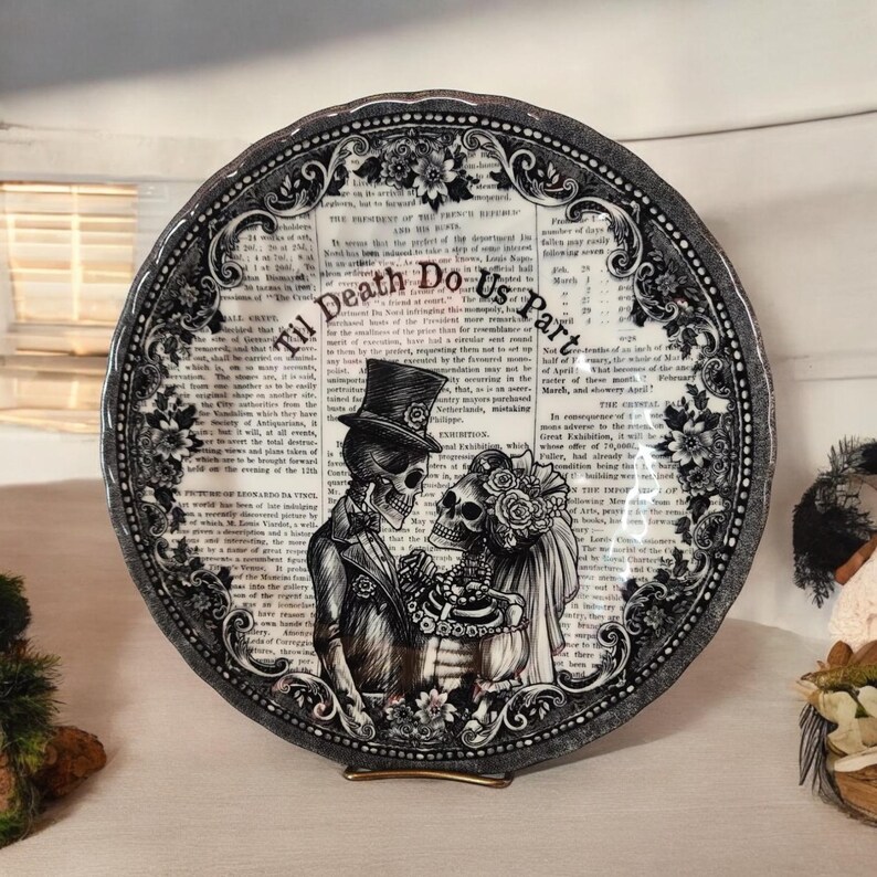 K&ouml;nnte beinhalten: Schwarzer und wei&szlig;er Keramikteller mit einem gotischen Hochzeitsthema. Der Teller zeigt ein Skelettpaar mit einem Blumendesign und dem Text "Til Death Do Us Part".