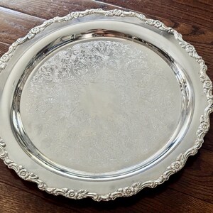 May include: A round silver-toned serving tray featuring a polished center with an intricate floral and scrollwork engraving. The rim has a raised, scalloped edge decorated with embossed flower motifs and a detailed border pattern.