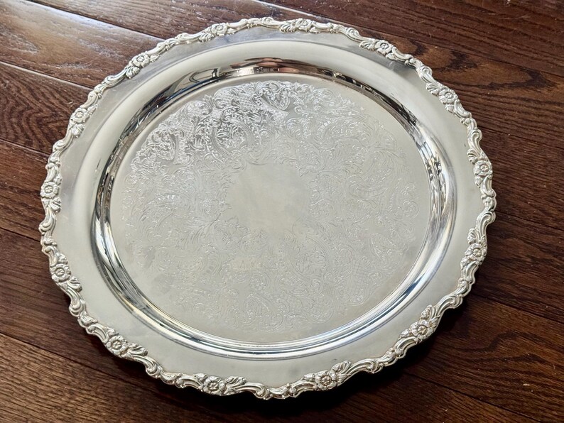 May include: A round silver-toned serving tray featuring a polished center with an intricate floral and scrollwork engraving. The rim has a raised, scalloped edge decorated with embossed flower motifs and a detailed border pattern.