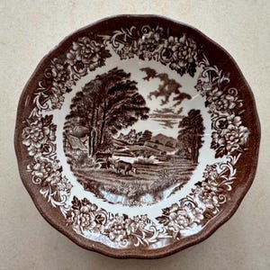 May include: A round ceramic bowl with a scalloped edge, featuring a brown and white transferware pattern. The center depicts a pastoral landscape with trees, cattle, and buildings, while the rim is decorated with a dense floral border.