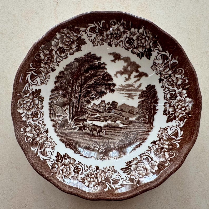 May include: A round ceramic bowl with a scalloped edge, featuring a brown and white transferware pattern. The center depicts a pastoral landscape with trees, cattle, and buildings, while the rim is decorated with a dense floral border.