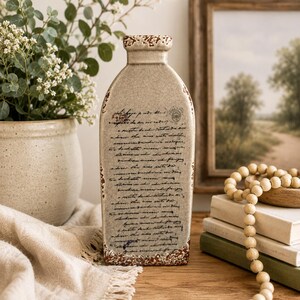 May include: A rectangular, beige ceramic vase with a distressed, crackled finish. The vase features handwritten script and decorative brown edging. A neutral-toned vase for home decor.