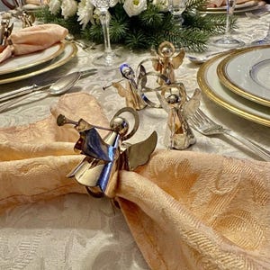 K&ouml;nnte beinhalten: Festliche Tischdekoration mit goldenen Tellern, Kristallgl&auml;sern und Engeln als Serviettenringen. Ein Blumengesteck mit wei&szlig;en Blumen und Gr&uuml;nzeug sorgt f&uuml;r einen nat&uuml;rlichen Akzent. Pfirsichfarbene Servietten schaffen eine warme und einladende Atmosph&auml;re.