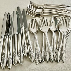 May include: A collection of polished silver flatware, including knives, forks, and spoons. The handles feature a delicate floral design. The silverware is arranged on a light-colored surface, showcasing the reflective quality of the metal.