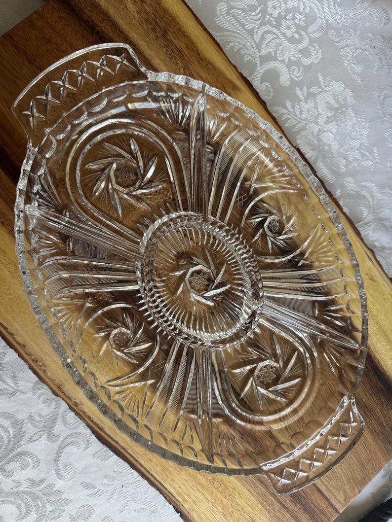 Op de afbeelding: Transparante glazen serveerschaal met een ovale vorm en een stervormig ontwerp in het midden. De schaal heeft een verdeeld ontwerp met decoratieve handvatten aan elk uiteinde. Het glas heeft een geslepen kristal uiterlijk.