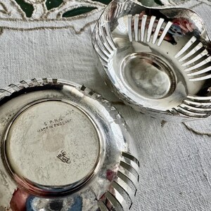 May include: Two silver-colored, decorative bowls. One bowl is upside down, revealing a circular base with the text "E.P.N.S. MADE IN ENGLAND". The other bowl has a slotted design. Both bowls are on a textured, off-white surface.