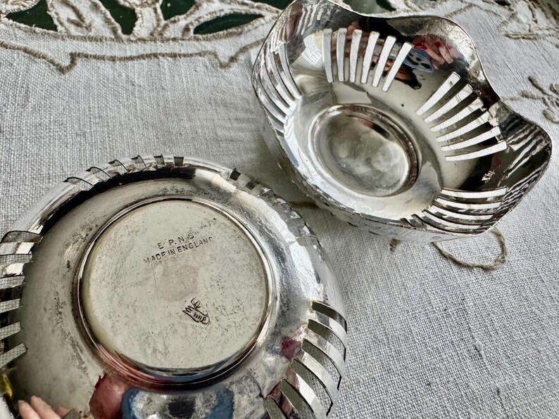 May include: Two silver-colored, decorative bowls. One bowl is upside down, revealing a circular base with the text "E.P.N.S. MADE IN ENGLAND". The other bowl has a slotted design. Both bowls are on a textured, off-white surface.