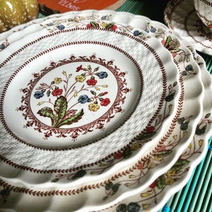 Op de afbeelding: Een set van vier witte keramische borden met een bruin, rood en blauw bloemenpatroon. De borden hebben een geweven ontwerp en gegolfde randen.