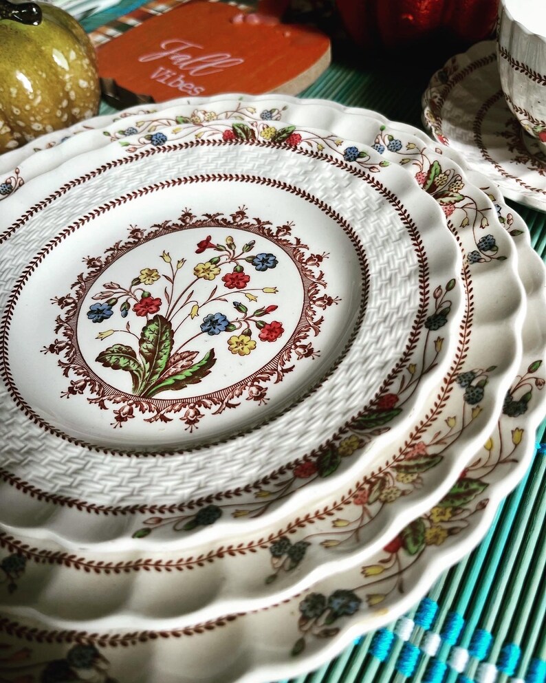 Op de afbeelding: Een set van vier witte keramische borden met een bruin, rood en blauw bloemenpatroon. De borden hebben een geweven ontwerp en gegolfde randen.